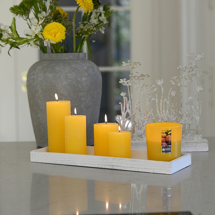 Zwei brennende weiße Stumpenkerzen vom Hersteller Müller auf einem weißen Untergrund. Neben den Kerzen liegen zwei rosa Tulpen mit grünen Blättern.