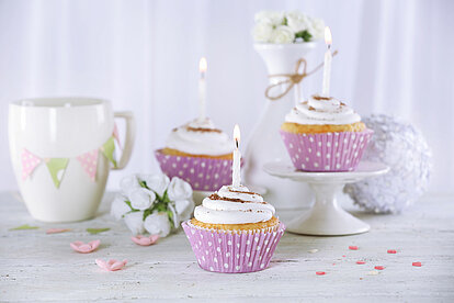 Ein liebevoll dekorierter Geburtstagstisch mit drei Cupcakes in rosa Bechern, auf denen jeweils eine weiße Geburtstagskerze brennt.