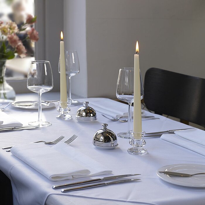 Vier brennende weiße Stabkerzen in zwei verschiedenen Haltern, die auf einem hellen Tisch stehen. Neben den Kerzen befinden sich ein Glas-Krug und zwei leere Gläser. Im Vordergrund liegt ein Teller mit goldenem Besteck. Im leicht verschwommenen Hintergrun