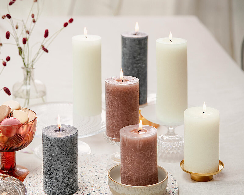 Auf einem weißen Tisch steht am linken Bildrand eine Schüssel mit leckeren bunten Macarons sowie ein Stapel gläserner Dessertteller. In der Mitte des Bildes ist eine runde Steinplatte auf der Steinplatte neben und hinter der Steinplatte stehen mehrere brennende Stumpenkerzen in den Farben weiche Perle, Wildleder Braun und stürmisches Grau von Bolsius.