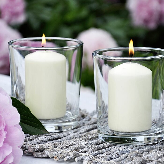 Eine brennende rosa Müller-Kerze in einem Glaswindlicht, platziert in einem weißen geflochtenen Korb. Im Hintergrund steht ein Strauß aus rosa Rosen und weißen Schleierkraut-Blüten in einer klaren Vase auf einem Tisch. Die Kombination verleiht dem Bild ei