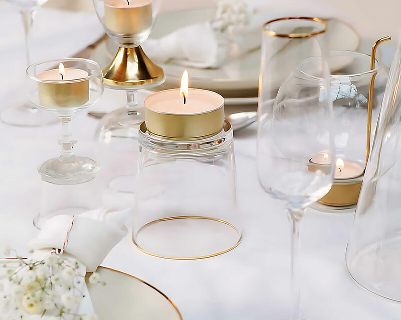 Ein festlich gedeckter Tisch mit weissen und goldenen Elementen wie Tellern, Gläsern, Messern und Gabeln sowie mehrere Teelichthalter auf denen sich brennende Teelichter von Bolsius in einem goldenen Becher befinden. Die Dekoration verleiht dem Bild eine Atmosphäre wie auf einer festlich gedeckten Hochzeitstafel.