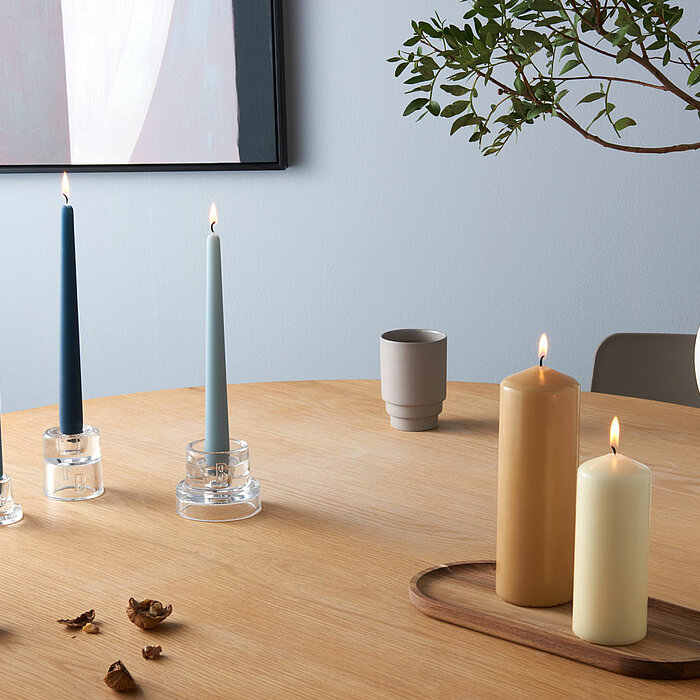 Zwei brennende Bolsius Stumpenkerzen in den Farben Beige und Weiß stehen auf einem hölzernen Tablett auf einem runden Holztisch. Daneben sind drei Bolsius Leuchterkerzen in verschiedenen Blautönen auf gläsernen Kerzenhaltern angezündet. Der Tisch ist mit 