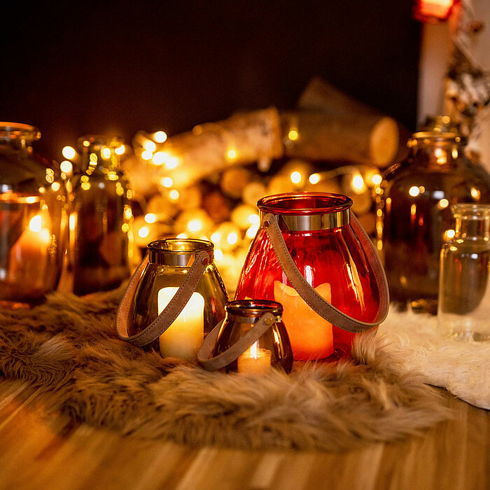 Mehrere Sandra Rich Laternen und Glasbehälter in verschiedenen Größen und Farben sind um einen gemütlich beleuchteten Kaminsims angeordnet. Die Laternen, unter anderem in leuchtendem Rot und warmem Orange, befinden sich auf weichen, pelzigen Teppichen, um