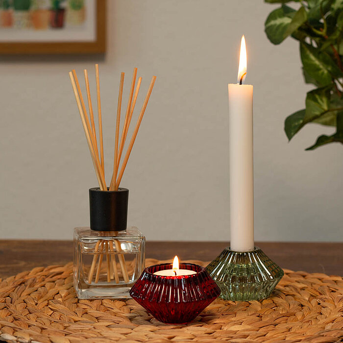 Eine brennende weiße Stabkerze in einem grünen Glas-Kerzenhalter, eine brennende rote Teelichtkerze in einem rubinroten Glas-Teelichthalter und ein Duftdiffusor mit Holzstäbchen in einem klaren Glasbehälter von Edelman stehen auf einem runden, geflochtene