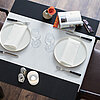Ein elegant gedeckter Tisch mit Produkten von Duni. Auf einem schwarzen Tisch liegen ein weißer Tischläufer und zwei weiße Teller mit darauf platzierten weißen Stoffservietten. Jeder Platz ist mit einem schwarzen Besteckset und zwei Weingläsern versehen. 