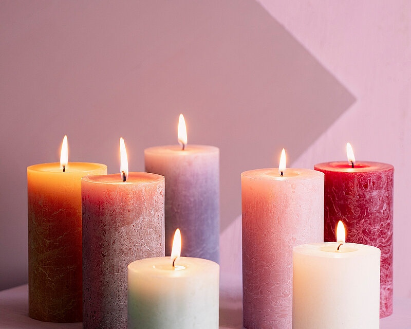 Vor einer zarten, altrosafarbenen Wand stehen mehrere rustikale, unterschiedlich hohe und brennende Stumpenkerzen von Bolsius in verschiedenen zarten Pastell- und Rottönen, hintereinander verteilt. Die Szene strahlt eine warme, beruhigende Atmosphäre aus, und zeigt ein kleiner Teil der farblichen Vielfalt der Bolsius Kerzen.