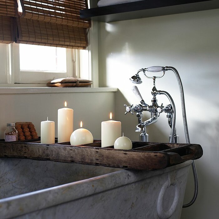 Fünf brennende Bolsius Stumpenkerzen in unterschiedlichen Größen und Formen, darunter zwei Kugelkerzen, stehen auf einem hölzernen Tablett, das quer über einer antik aussehenden Badewanne platziert ist. Im Hintergrund befinden sich Handtücher, ein Fenster