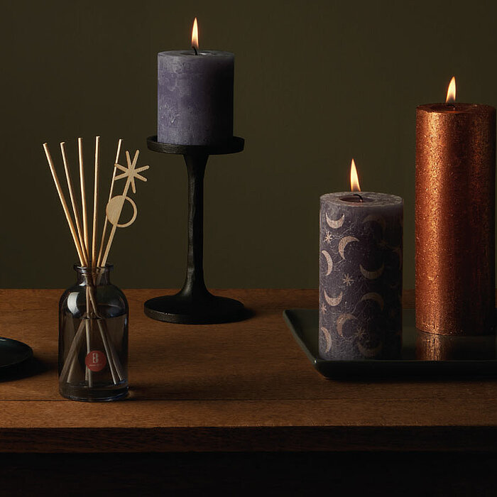 Vier brennende Kerzen in den Farben Lila, Dunkelviolett, Rostrot und Koralle stehen auf einer Holzfläche in einer harmonischen Anordnung. Die Kerzen haben unterschiedliche Muster und Texturen und sind auf schwarzen Kerzenhaltern platziert. Links von der M