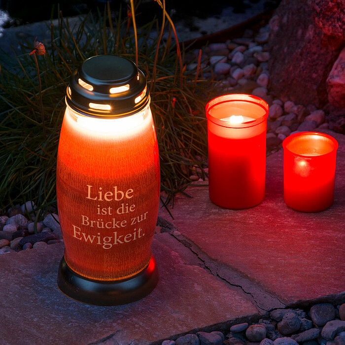 Eine große rote Grabkerze mit der Aufschrift "Liebe ist die Brücke zur Ewigkeit" steht neben zwei kleineren brennenden roten Kerzen auf einem gepflasterten Weg, der von Kieselsteinen und etwas Gras umgeben ist. Die Szene findet im Freien statt und wird vo