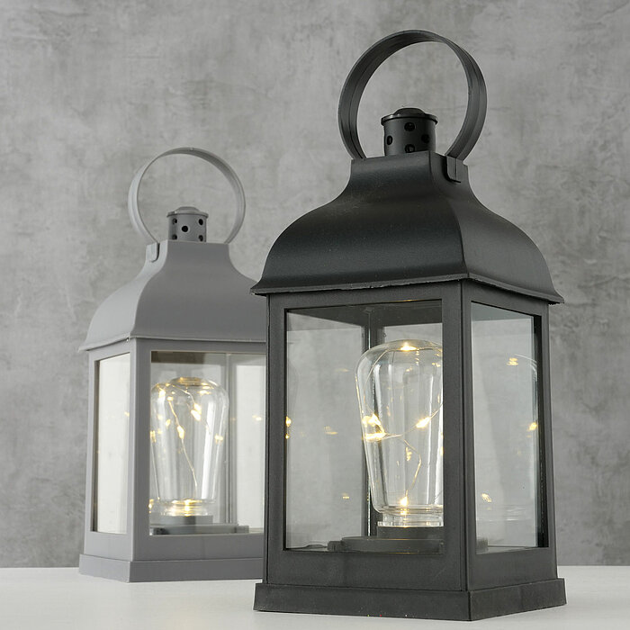 Zwei Laternen stehen auf einer weißen Oberfläche vor einem grauen Hintergrund. Die vordere Laterne ist schwarz und die hintere Laterne ist grau. Beide Laternen haben transparente Wände und enthalten eine Glühbirne mit kleinen Lichterketten darin, die ein 
