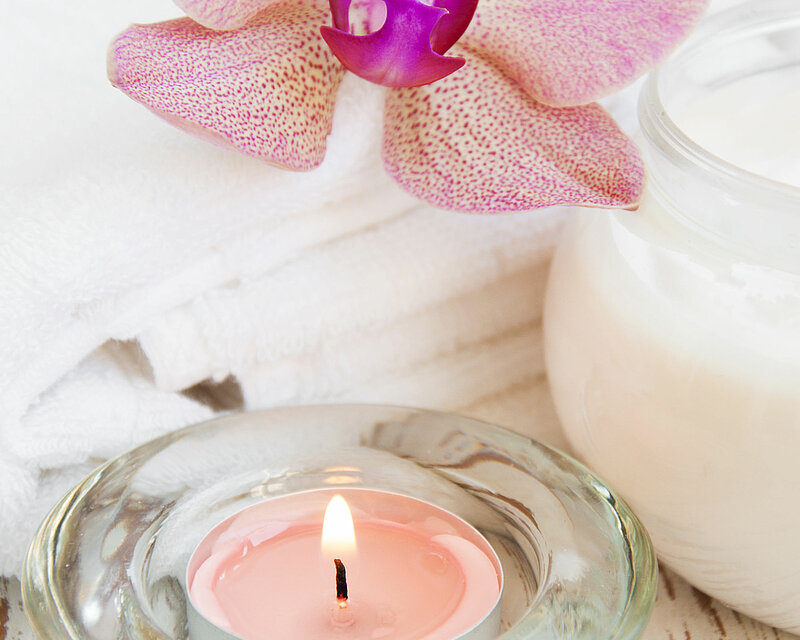Auf einem weißen, leicht marmorierten Tisch steht ein Teelicht mit dem Duft "Englische Rose" in einem transparenten Glas-Teelichthalter von Müller-Kerzenfabrik. Am rechten Bildrand ist zur Hälfte ein bauchiges Glasgefäß zu sehen, das mit weißem Wachs gefüllt ist. Über dem Arrangement thront eine rosafarbene Orchideenblüte. Das gesamte Bild strahlt Helligkeit und Frieden aus und verbreitet eine angenehme, entspannte Stimmung.