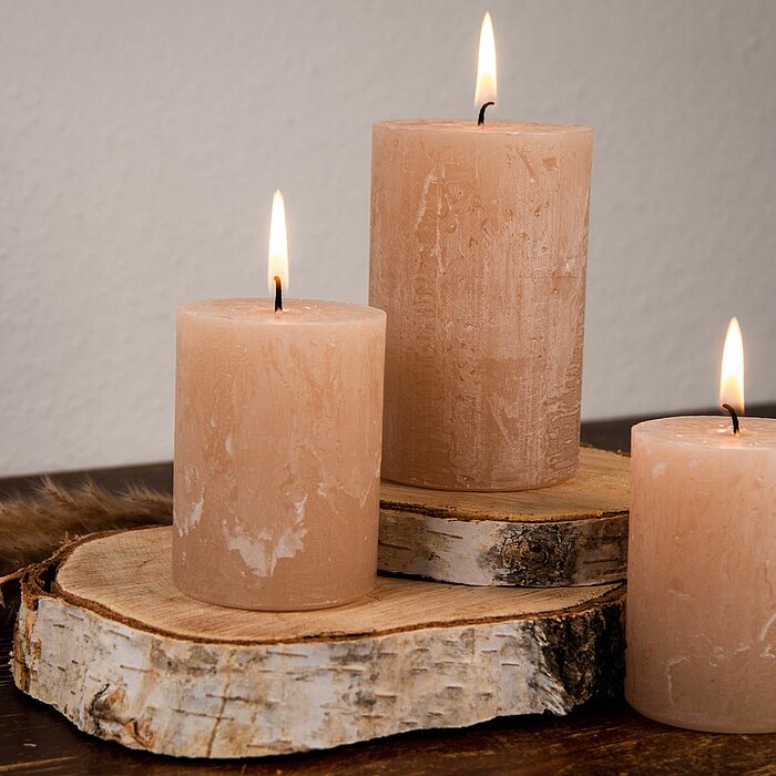 Drei brennende DecoLite-Stumpenkerzen in verschiedenen Höhen stehen auf zwei Holzscheiben, die als Kerzenständer dienen. Die Kerzen haben eine natürliche, beige Farbe und befinden sich auf einer dunklen Holzoberfläche. Im Hintergrund ist eine schlichte Wa