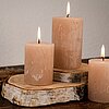 Drei brennende DecoLite-Stumpenkerzen in verschiedenen Höhen stehen auf zwei Holzscheiben, die als Kerzenständer dienen. Die Kerzen haben eine natürliche, beige Farbe und befinden sich auf einer dunklen Holzoberfläche. Im Hintergrund ist eine schlichte Wa