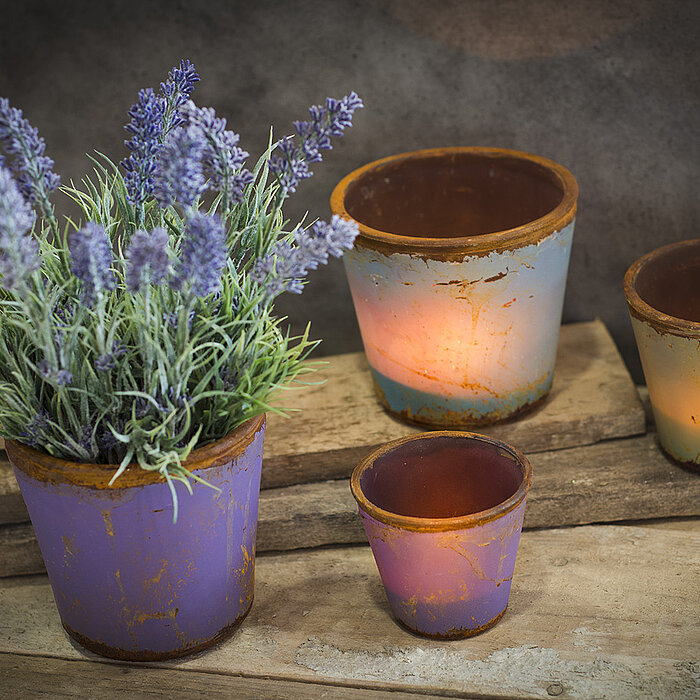 Vier rustikal anmutende Kerzenhalter von Sandra Rich in verschiedenen Größen stehen auf einer Holzoberfläche. Drei davon sind mit brennenden Teelichtern beleuchtet, die ein warmes Licht ausstrahlen. Der größte Kerzenhalter ist mit einem Lavendelstrauß gef