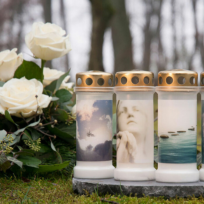 Vier Grabkerzen von Bolsius mit goldenen Deckeln stehen nebeneinander auf einem Stein. Jede Kerze ist mit einem anderen Bild verziert: eine betende Person, eine weiße Blume, fliegende Vögel und eine Pusteblume. Links neben den Kerzen liegt ein Strauß weiß