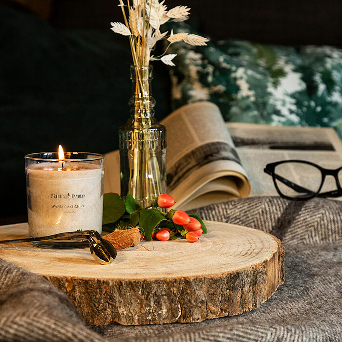 Eine brennende Duftkerze von Prices Candles auf einer runden Holzscheibe, daneben liegen eine goldfarbene Kerzenschere und einige rote Beeren. Im Hintergrund sind eine geöffnete Zeitung, eine Brille, ein grau-grünes Kissen und eine Vase mit getrockneten B
