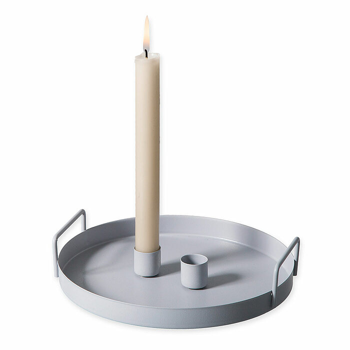 Eine brennende Stabkerze von Halbach in einem hellen Kerzenhalter, der auf einem runden silberfarbenen Tablett mit Griffen steht. Das schlichte, minimalistische Design des Kerzenhalters und Tabletts vermittelt eine ruhige und moderne Stimmung.