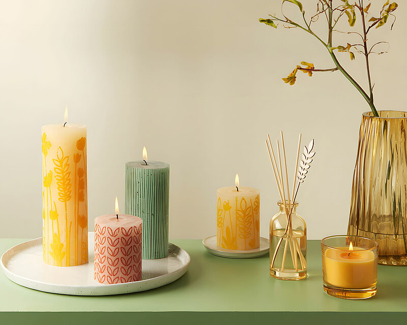 Sommerliche Kombination aus Rustik Stumpenkerzen mit Motiv in den Farben Sonnengelb, Türkis und Rosa. Die Kerzen stehen auf einem weißen Tablett, neben einem Raumduft Diffuser und einer gelben Duftkerze im Glas.