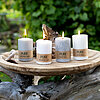 Vier brennende Kerzen vom Hersteller "Wenzel" stehen auf einer runden Holzplatte, die auf einem verwitterten Baumstamm positioniert ist. Die Kerzen sind in verschiedenen Weiß- und Grautönen gehalten und tragen die Aufschrift "PURE NATURAL WAX" und "PURE O