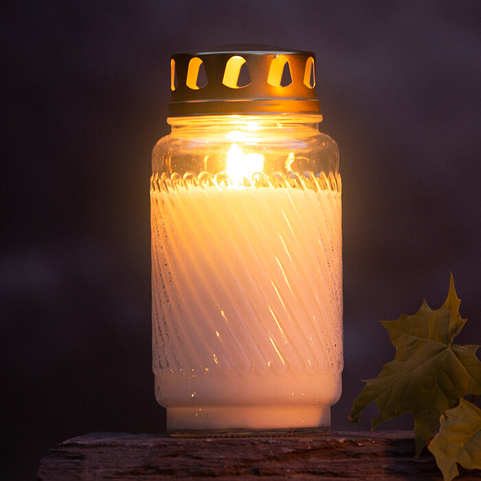 Eine brennende Bolsius Grabkerze in einem gläsernen Behälter mit einem goldenen Deckel. Die Kerze steht auf einem Stein neben einigen grünen Blättern. Der Hintergrund ist dunkel und hebt die warme, ruhige Atmosphäre der leuchtenden Kerze hervor.