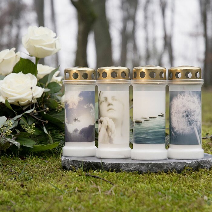 Vier Bolsius Grabkerzen mit weißen Wachs in durchsichtigen Glasbehältern stehen nebeneinander auf einer dunklen Steinplatte. Jede Kerze hat einen goldenen Deckel mit Löchern und ist mit verschiedenen beruhigenden Natur- und Seelenbildern bedruckt, darunte