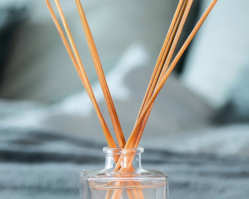 Ein Prices Raumduft-Diffuser mit Rattan-Stäbchen steht auf einem floral dekorierten Tablett. Im Hintergrund und unter dem Tablett ist eine klare Stoffdecke zu sehen. Die Szene strahlt eine ruhige Eleganz aus und lädt dazu ein, sich inmitten des angenehmen Dufts zu entspannen.