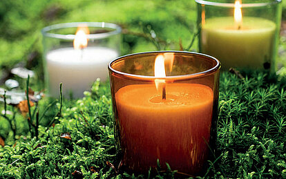 Nahaufnahme einer brennenden Stearin Kerze in einem orangefarbenen Glas. Im Hintergrund erkennt man noch zwei weitere Kerzen, eine mit grünem Glas und eine mit klarem. Die Gläser stehen auf dichtem, grünem Moos.