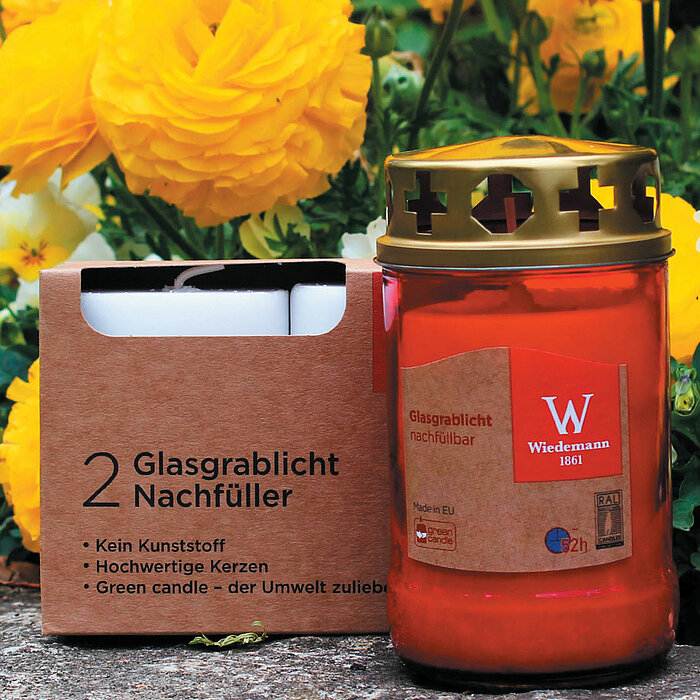 Ein rotes Grablicht von Wiedemann mit einem goldenen Deckel steht auf einem Stein. Auf dem Etikett steht "Glasgrablicht nachfüllbar" und es ist in der EU hergestellt. Das Grablicht steht im Vordergrund, während im Hintergrund ein unscharfer Friedhofsberei