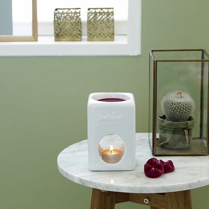 Eine Bolsius-Duftkerze brennt in einem weißen Kerzenhalter mit einem offenen Design, durch den der Kerzenschein sichtbar ist. Auf dem Holztisch daneben liegen rosa Blüten und ein weiteres Duftkerzen-Accessoire. Im Hintergrund sitzt eine Person gemütlich i