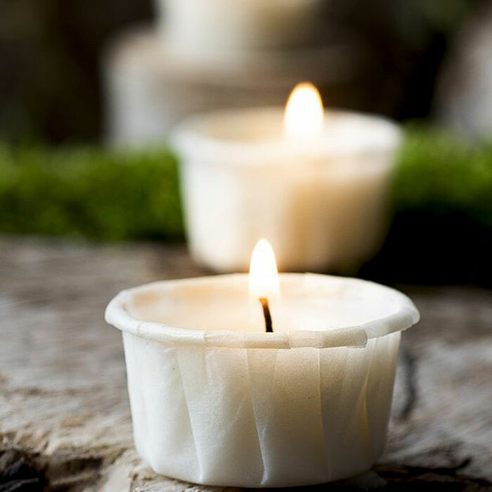 Drei weiße, brennende Teelichter stehen auf einer natürlichen, rustikalen Holzoberfläche. Im Hintergrund ist eine grüne, unscharfe Pflanze zu erkennen. Die Kerzen strahlen ein warmes, beruhigendes Licht aus und verleihen dem Bild eine gemütliche und ruhig