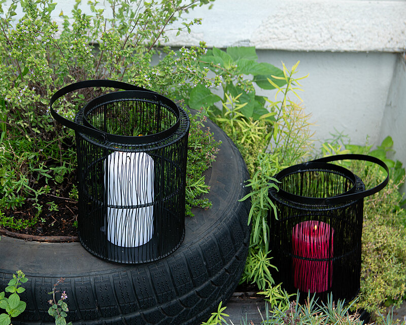 auf dem bild befinden sich zwei moderne Laternen Yves von Edelman. Eine davon steht auf der linken Seite erhöht auf einem mit grünen frischen Pflanzen bepflanzten Autoreifen. Die zweite Laterne steht auf dem Boden neben frischen und freundlichen Pflanzen und grünen Gräsern. In beiden Laternen befindet sich je eine weisse und eine rote brennende Stumpenkerze.