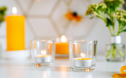 Im Vordergrund steht ein brennendes, gelbes Duft Teelicht in einem Glashalter. Im Hintergrund sieht man weitere gelbe Kerzen brennen, sowie eine Vase mit Blumen.