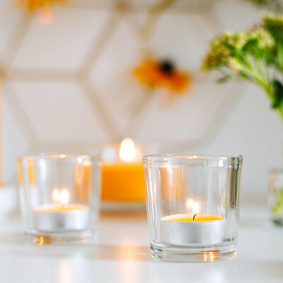 Im Vordergrund steht ein brennendes, gelbes Duft Teelicht in einem Glashalter. Im Hintergrund sieht man weitere gelbe Kerzen brennen, sowie eine Vase mit Blumen.