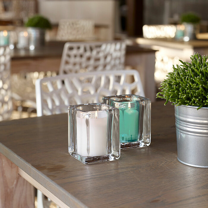 Zwei brennende Bolsius Kerzen in quadratischen Glasbehältern, eine in Weiß und eine in einem türkisen Farbton, stehen auf einem Holztisch. Daneben befindet sich eine silberne Metalltopfpflanze. Im Hintergrund sind weitere Tische und Stühle in einem hellen