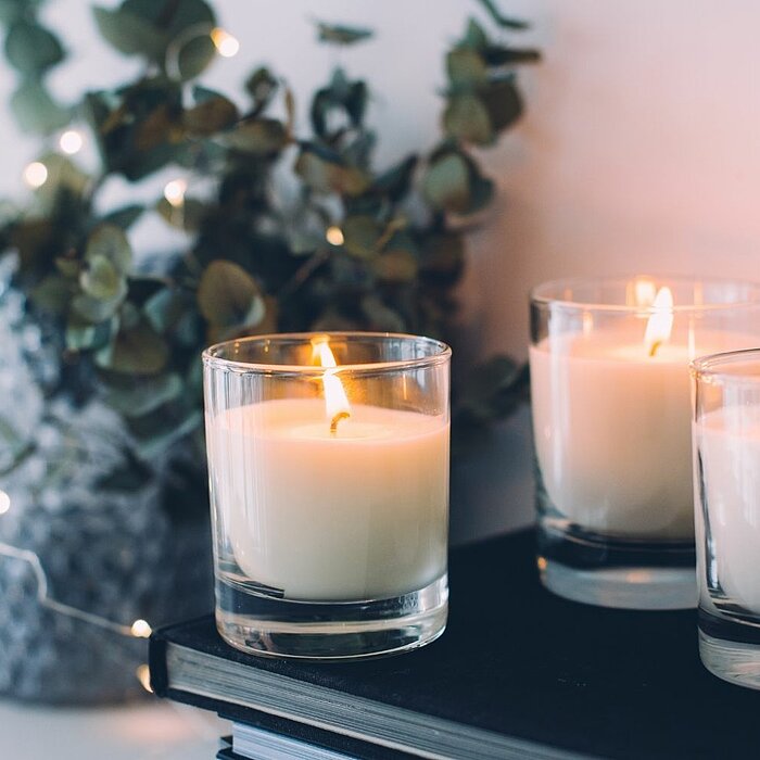 Drei brennende weiße Kerzen in transparenten Glasbehältern stehen nebeneinander auf einem dunklen Buch. Im Hintergrund ist eine Pflanze mit grünen Blättern in einem grauen Blumentopf zu sehen, umgeben von kleinen Lichterketten, die ein warmes und gemütlic
