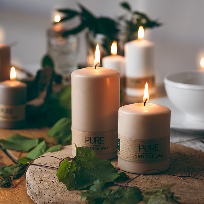 Neun brennende Kerzen des Herstellers Wenzel in verschiedenen Größen. Die Kerzen bestehen aus natürlichem Wachs und tragen das Label "PURE NATURAL WAX". Sie stehen auf einem Holz- und einem Steintablett, welches mit grünen Blättern dekoriert ist. Im Hinte