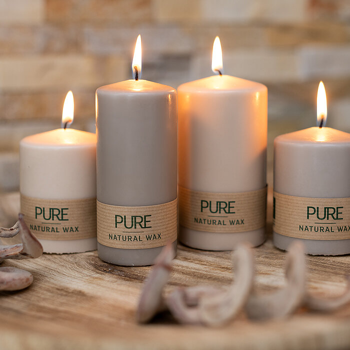 Vier brennende Kerzen aus natürlichem Wachs vom Hersteller "Wenzel" stehen auf einer hölzernen runden Platte. Die Kerzen tragen die Aufschrift "PURE NATURAL WAX" und sind in verschiedenen Größen angeordnet. Im Vordergrund liegen dekorative Holzstücke. Der