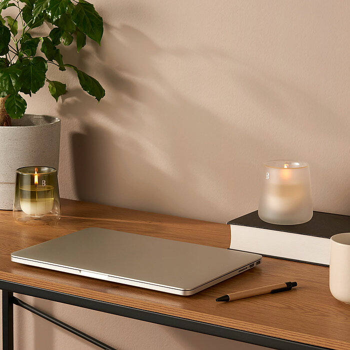 Ein aufgeräumter Schreibtisch mit einem minimalistischen und ruhigen Ambiente. Auf der Holzoberfläche des Schreibtisches befindet sich ein geschlossener Laptop, ein Stift und eine weiße Tasse. Daneben steht eine brennende, in einen matten Glasbehälter geh