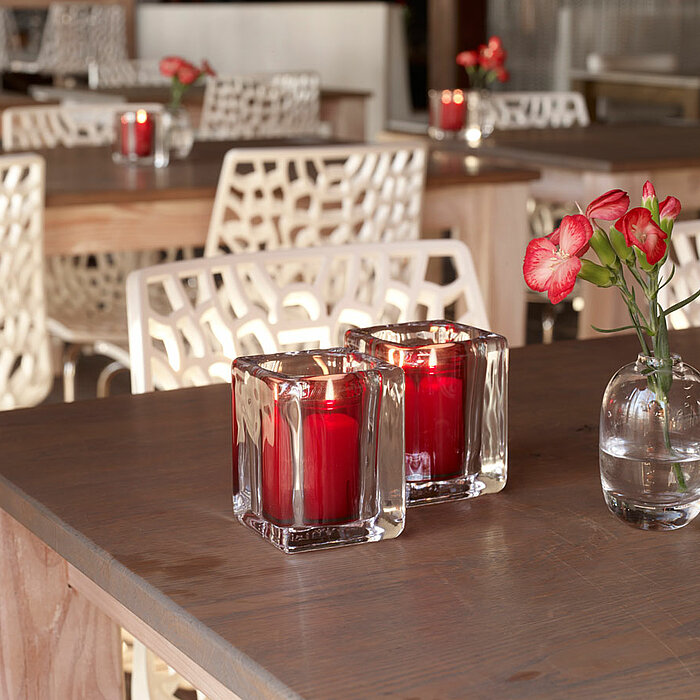 Zwei brennende rote Bolsius Kerzen in quadratischen Glasbehältern stehen nebeneinander auf einem Holztisch. Daneben befindet sich eine kleine Glasvase mit roten Blumen. Im Hintergrund sind Holz-Esstische mit weißen, geometrisch gemusterten Stühlen zu sehe