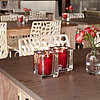 Zwei brennende rote Bolsius Kerzen in quadratischen Glasbehältern stehen nebeneinander auf einem Holztisch. Daneben befindet sich eine kleine Glasvase mit roten Blumen. Im Hintergrund sind Holz-Esstische mit weißen, geometrisch gemusterten Stühlen zu sehe