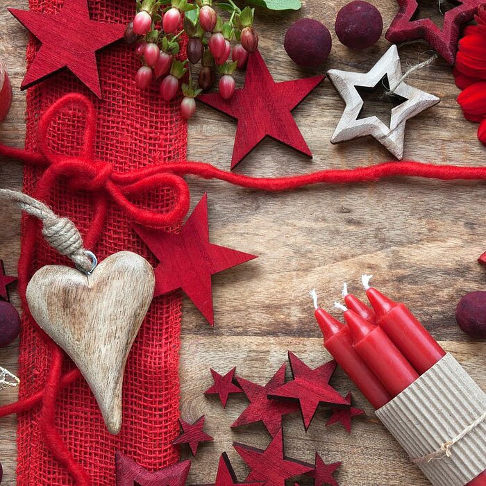 Vier rote Wenzel-Kerzen, die zu einem Bündel zusammengebunden sind, liegen auf einem hölzernen Untergrund. Neben ihnen befinden sich rote und weiße Sterne aus Holz, eine rote Stumpenkerze, dekorative Beeren und eine rote Blüte. Ein Herzanhänger aus Holz u