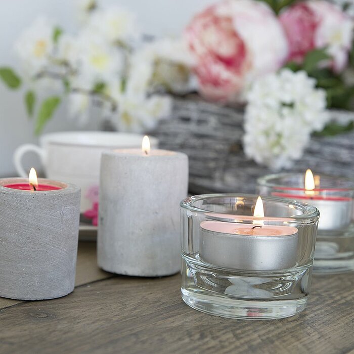 Drei brennende Teelichter vom Hersteller Müller, eines in Gelb, eines in Blau und eines in Türkis, befinden sich in klaren Glasbehältern auf einer weißen, strukturierten Holzoberfläche. Rund um die Glasbehälter liegen einige weiße Steine verteilt, was ein