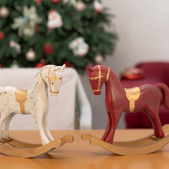 Zwei Edelman Schaukelpferde, eines in Weiß und das andere in Rot, jeweils mit goldenen Akzenten, stehen auf einem Holztisch. Im Hintergrund ist ein weihnachtlich geschmückter Baum mit Kugeln und eine rote Couch zu sehen. Die Schaukelpferde strahlen nostal