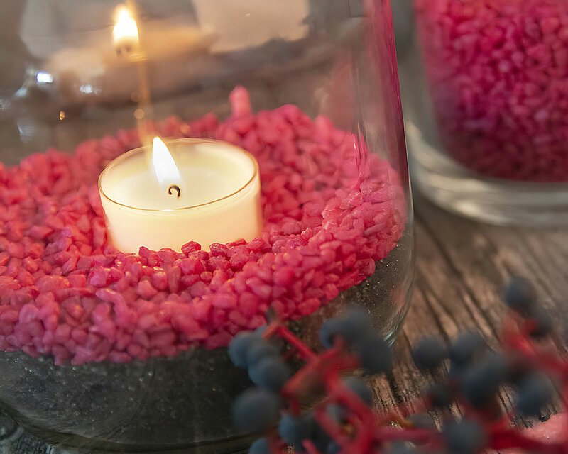 Auf einer rustikalen Tischplatte stehen mehrere Kerzengläser, gefüllt mit schwarzem Sand und roten Dekosteinen. In jedem Glas brennt ein Teelicht von Wenzel in einem klaren Becher. Der warme Schein der Kerzen erzeugt ein gemütliches und beruhigendes Licht. Im Vordergrund liegt ein Zweig mit saftigen Blaubeeren, der das Arrangement harmonisch ergänzt