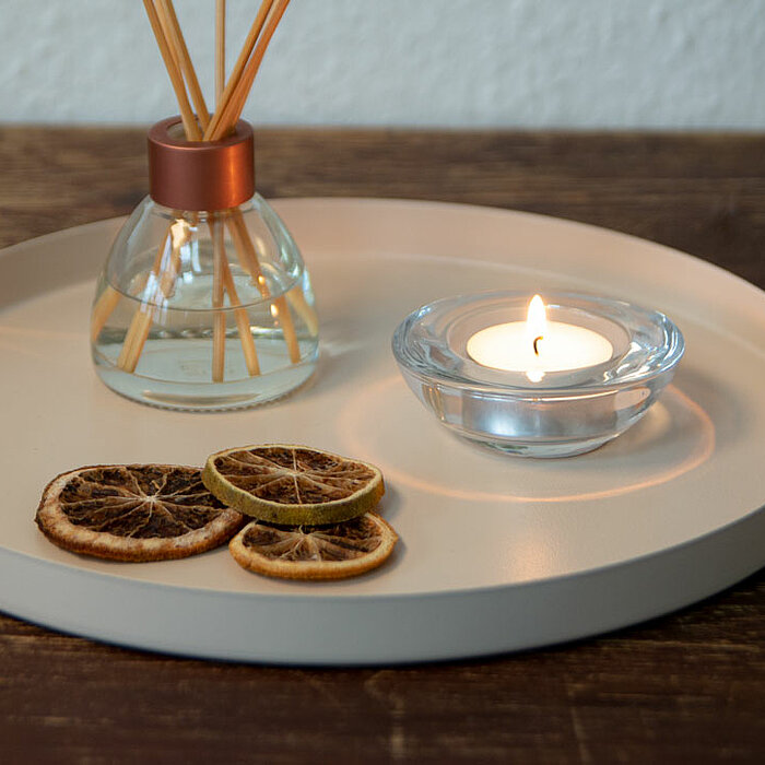 Auf einem runden Tablett mit braunen Lederriemen vom Hersteller "Edelman" stehen ein Glasgefäß mit Duftstäbchen und eine brennende Teelichtkerze in einem klaren Glashalter. Außerdem liegen mehrere getrocknete Zitronenscheiben auf dem Tablett, welches auf 