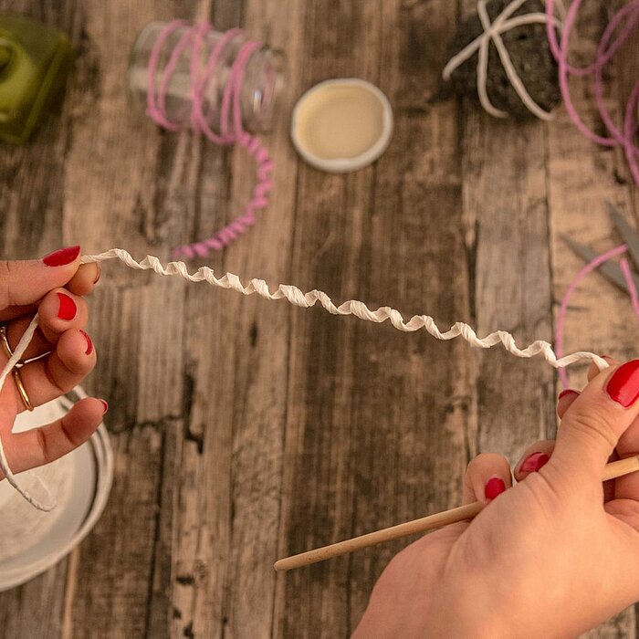 Ein dekoratives Arrangement von Krago: Eine brennende Teelichtkerze, umgeben von frischen Rosmarinzweigen und lila Kranzbändern, steht auf einer runden Holzscheibe. Daneben liegen zwei aufgeschlagene Bücher, die eine ruhige und besinnliche Atmosphäre scha