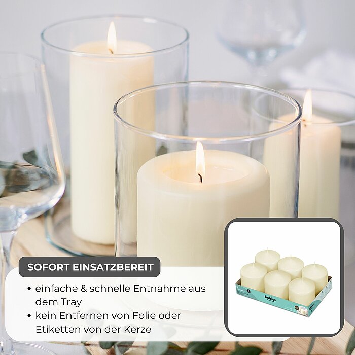 Sechs brennende Stumpenkerzen in verschiedenen Größen und Weißtönen stehen auf Glasuntersetzern auf einer schwarzen Bank. Im Hintergrund sind eine Treppe und ein aufgeschlagenes Buch auf einem weißen Tuch zu sehen. Der Raum wirkt ruhig und einladend.