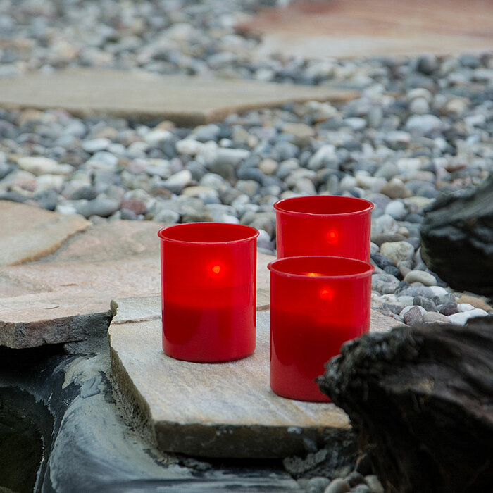 Drei brennende Bolsius Grabkerzen in roten Gläsern stehen auf einer Steinplatte. Sie sind umgeben von Kieselsteinen und großen Steinen, die eine ruhige und besinnliche Atmosphäre schaffen.