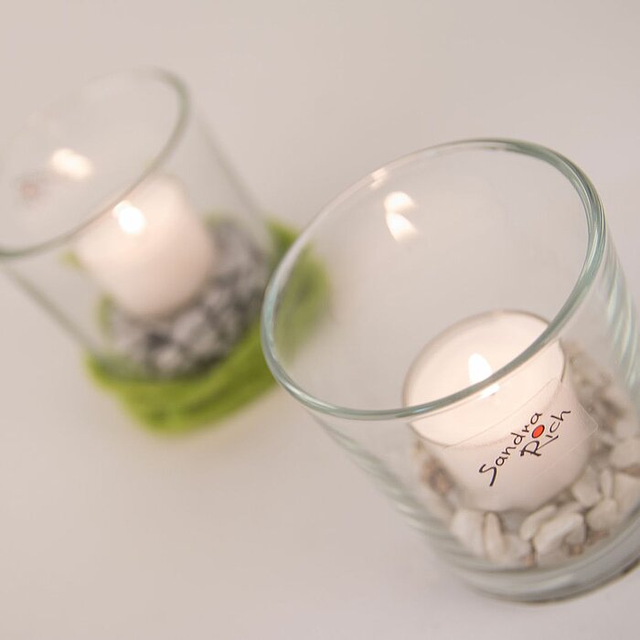 Zwei brennende Sandra Rich Teelichter in Glasbehältern, die auf weißen und grünen Dekosteinen ruhen. Die Glasbehälter sind schlicht und transparent gestaltet.